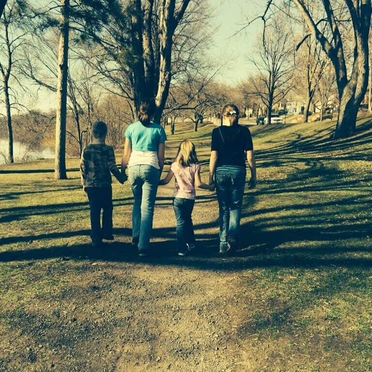 Two older children and two younger children walk hand-in-hand down a dirt path through a wooded park. The tall, leafless trees suggest early spring or late fall. Sunlight casts long shadows across the path, and a body of water with distant buildings appears in the background. The group is seen from behind, evoking a quiet moment of togetherness and reflection.
