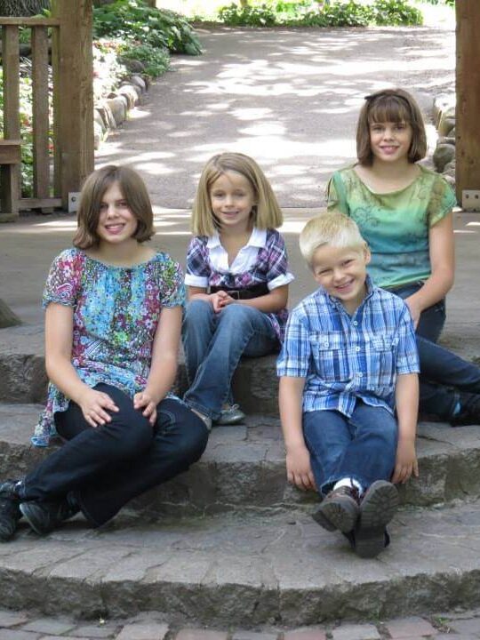 Four children sit together on stone steps in a garden or park, dressed casually and posed for a group photo. Behind them, a wooden structure with railings and lush greenery creates a peaceful outdoor setting. A paved path winds through the background. The timestamp “08/03/2013” marks the captured moment of togetherness and childhood connection.