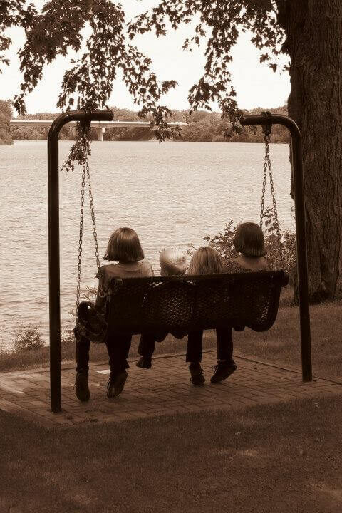 Three children sit together on a metal bench swing suspended by chains, overlooking a calm river. The swing rests on a paved area beside a grassy lawn, with trees surrounding the scene and a bridge visible in the distance. The sepia tone adds a nostalgic, timeless feel to this peaceful moment of companionship