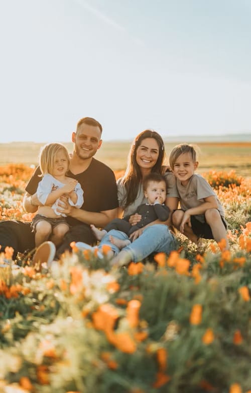 A family of five sits closely together in a field of vibrant orange flowers during golden hour. Warm sunlight bathes the scene, highlighting their smiles and the surrounding landscape. Behind them, open fields and distant hills stretch beneath a clear sky, creating a serene, joyful moment of connection and natural beauty.