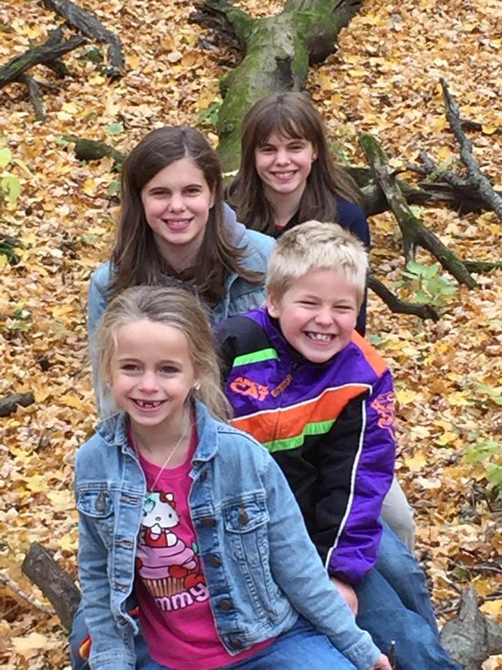 Four children sit in a line outdoors on a bed of fallen autumn leaves, smiling warmly at the camera. They’re dressed in casual, cool-weather clothing, surrounded by tree branches and a large fallen trunk in a forested setting. The scene captures a joyful, seasonal moment of togetherness and childhood connection.