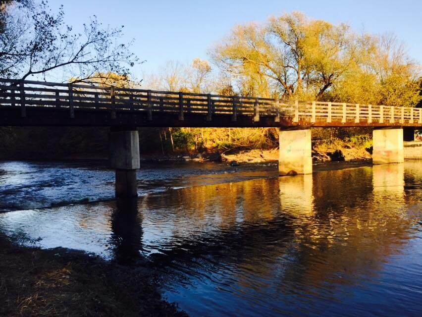 A wooden bridge with railings stretches across a calm river, supported by concrete pillars. The water reflects the bridge and surrounding trees, which glow with warm sunlight. Autumn-colored leaves and long shadows suggest early morning or late afternoon, creating a peaceful, seasonal atmosphere.