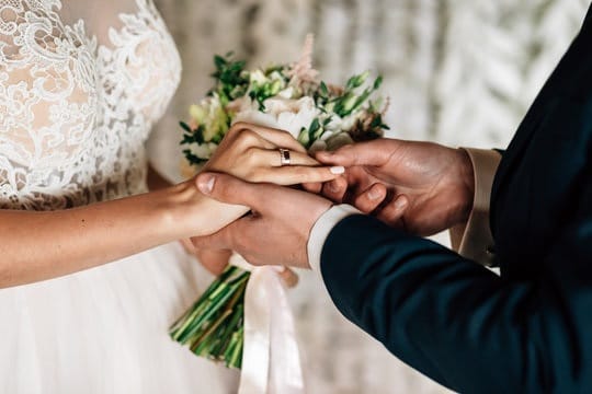 Close-up of a wedding ceremony moment where a person in a dark suit places a ring on the finger of a person wearing a white lace wedding dress and holding a bouquet. The focus is on their hands, capturing the exchange of rings—a symbol of love and commitment. The lace detail and flowers add elegance to this intimate, emotional scene.