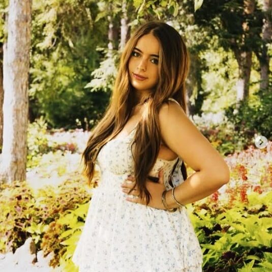 A person stands in a sunlit forest wearing a white, sleeveless, floral dress. Surrounded by tall trees and vibrant foliage in shades of green, yellow, and red, they pose confidently with one hand on their hip, looking toward the camera. The natural light and colorful setting create a serene, radiant atmosphere.